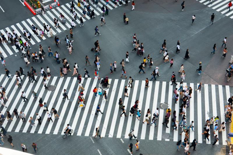 Overhead view of a busy city intersection with wide white zebra crosswalks filled with dozens of pedestrians walking in different directions across the asphalt. People carry bags and backpacks, and some pull rolling suitcases, creating a dynamic crisscross pattern against the bold striped crossings.