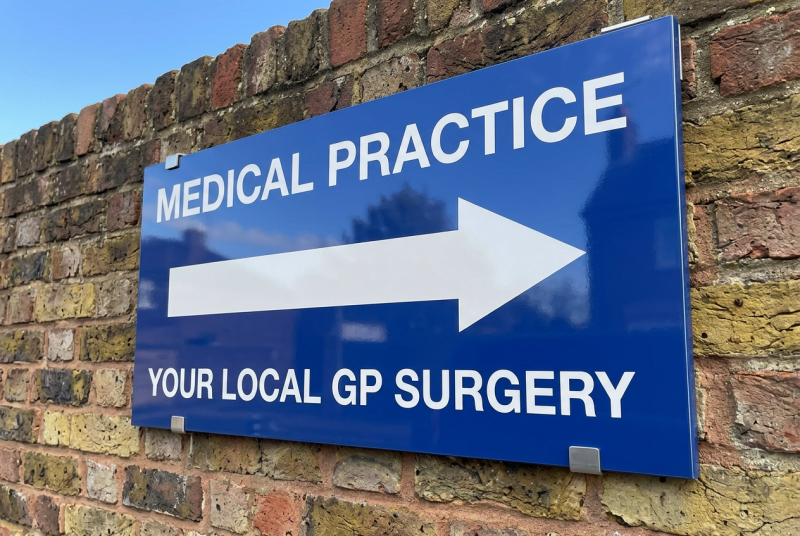 Blue rectangular sign mounted on a brick wall, with a large white left-pointing arrow above the white text ‘your local GP Surgery’