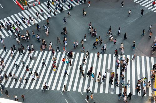 Overhead view of a busy city intersection with wide white zebra crosswalks filled with dozens of pedestrians walking in different directions across the asphalt. People carry bags and backpacks, and some pull rolling suitcases, creating a dynamic crisscross pattern against the bold striped crossings.